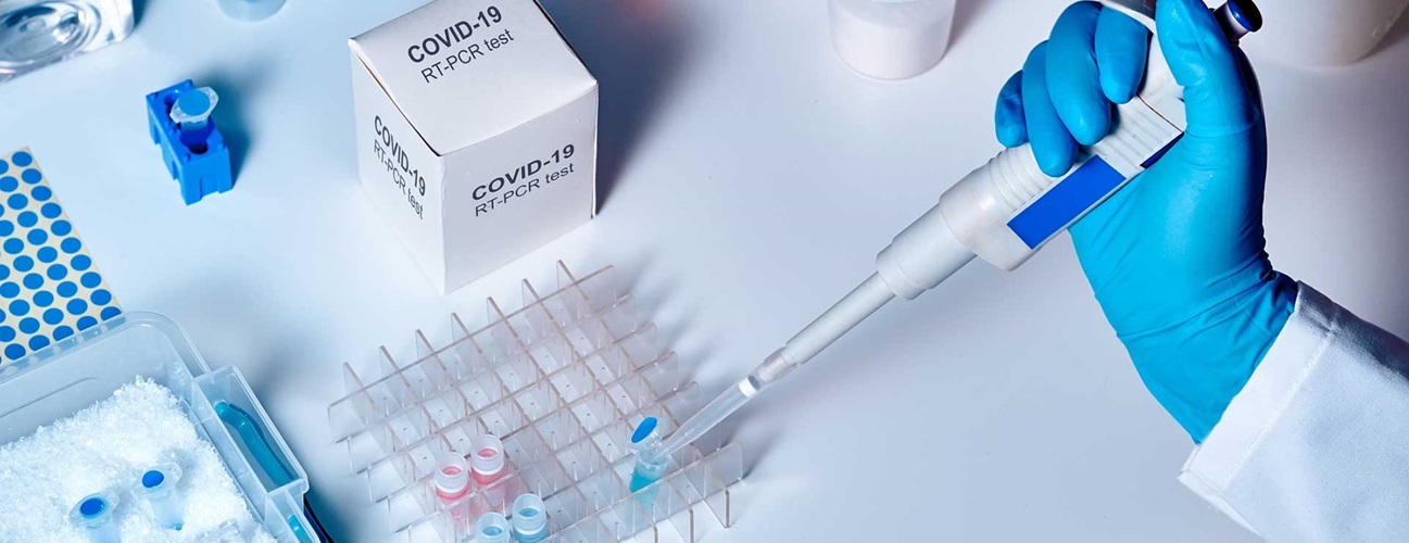 Ръка с ръкавица държи пипета, близо до кутия "COVID-19 RT-PCR тест" и лабораторно оборудване.